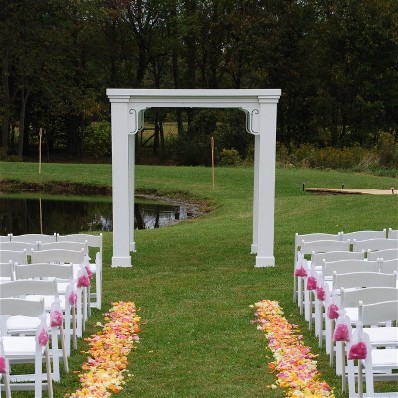 A 7' square arch appears at the end of an aisle lined with orange and pink rose petals. On either side of the aisle, rows of white garden chairs with pink bows are visible.