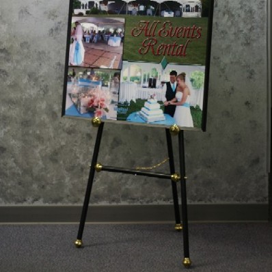 A simple black easel with gold accents holds an oversized poster with multiple wedding photos.